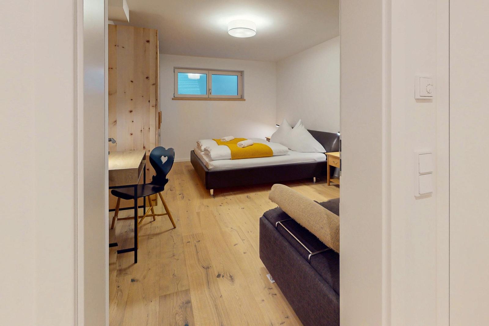 Schlafzimmer mit Bett, Schreibtisch und Holzboden. Fenster und Kleiderschrank sind sichtbar.