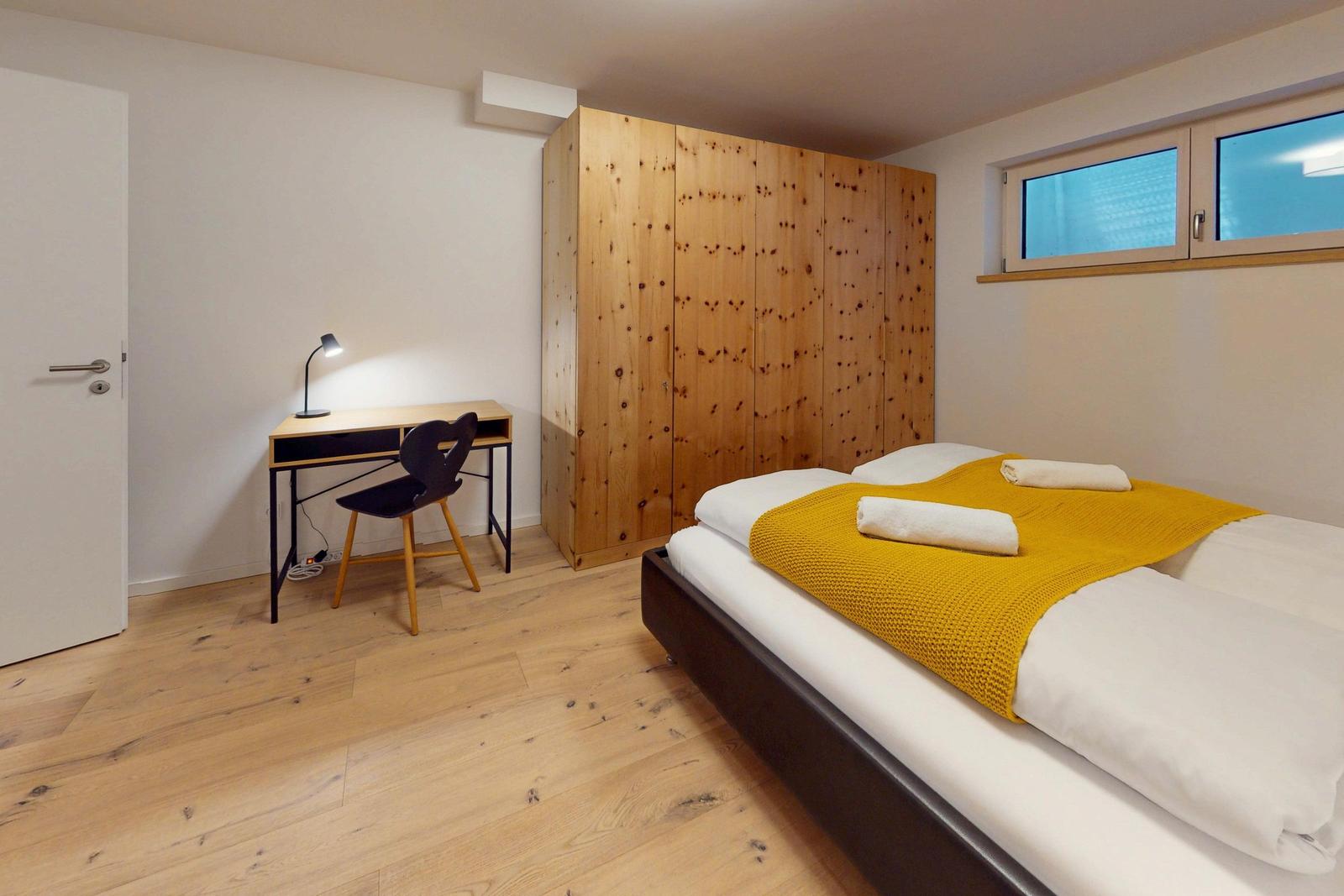 Schlafzimmer mit Doppelbett, Holzschrank und Schreibtisch mit Stuhl.