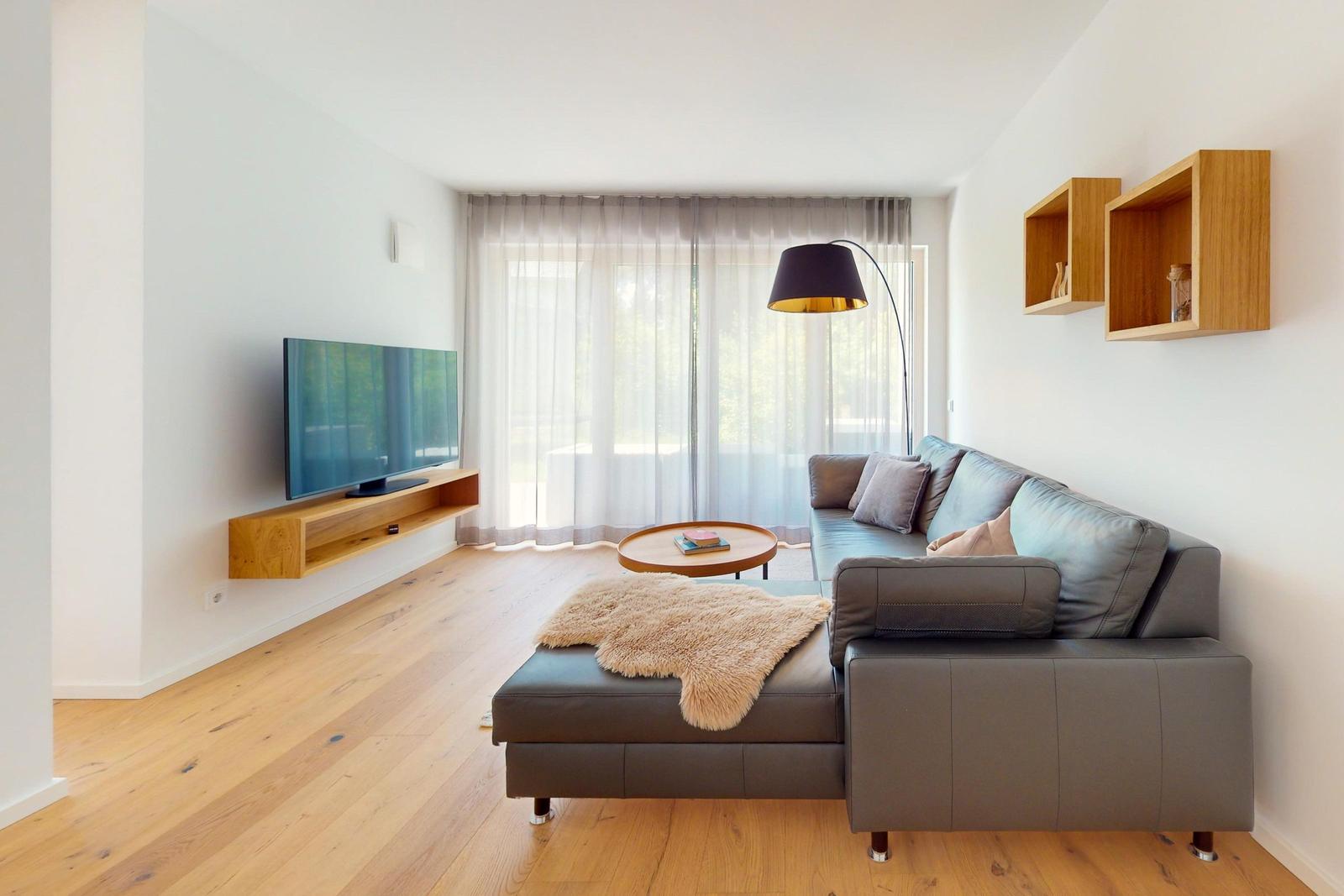 Wohnzimmer mit Sofa, Fernseher und Holzboden. Große Fenster mit Vorhängen.