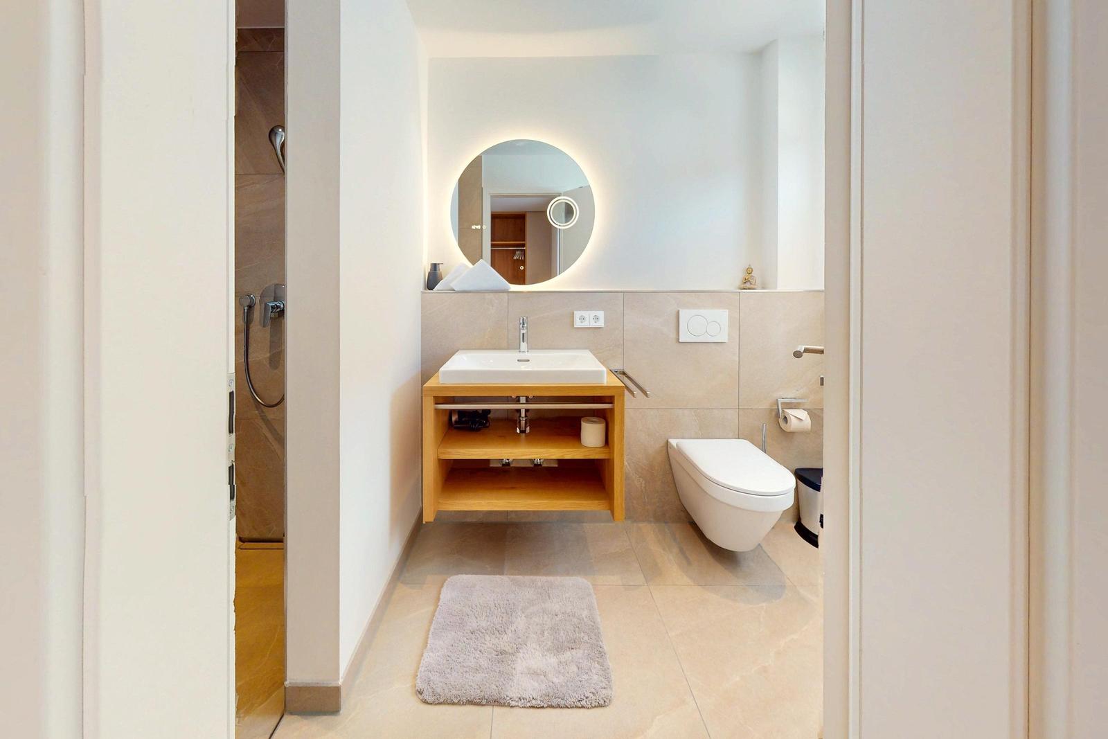 Badezimmer mit Waschbecken, WC und Dusche. Runder Spiegel mit Beleuchtung.