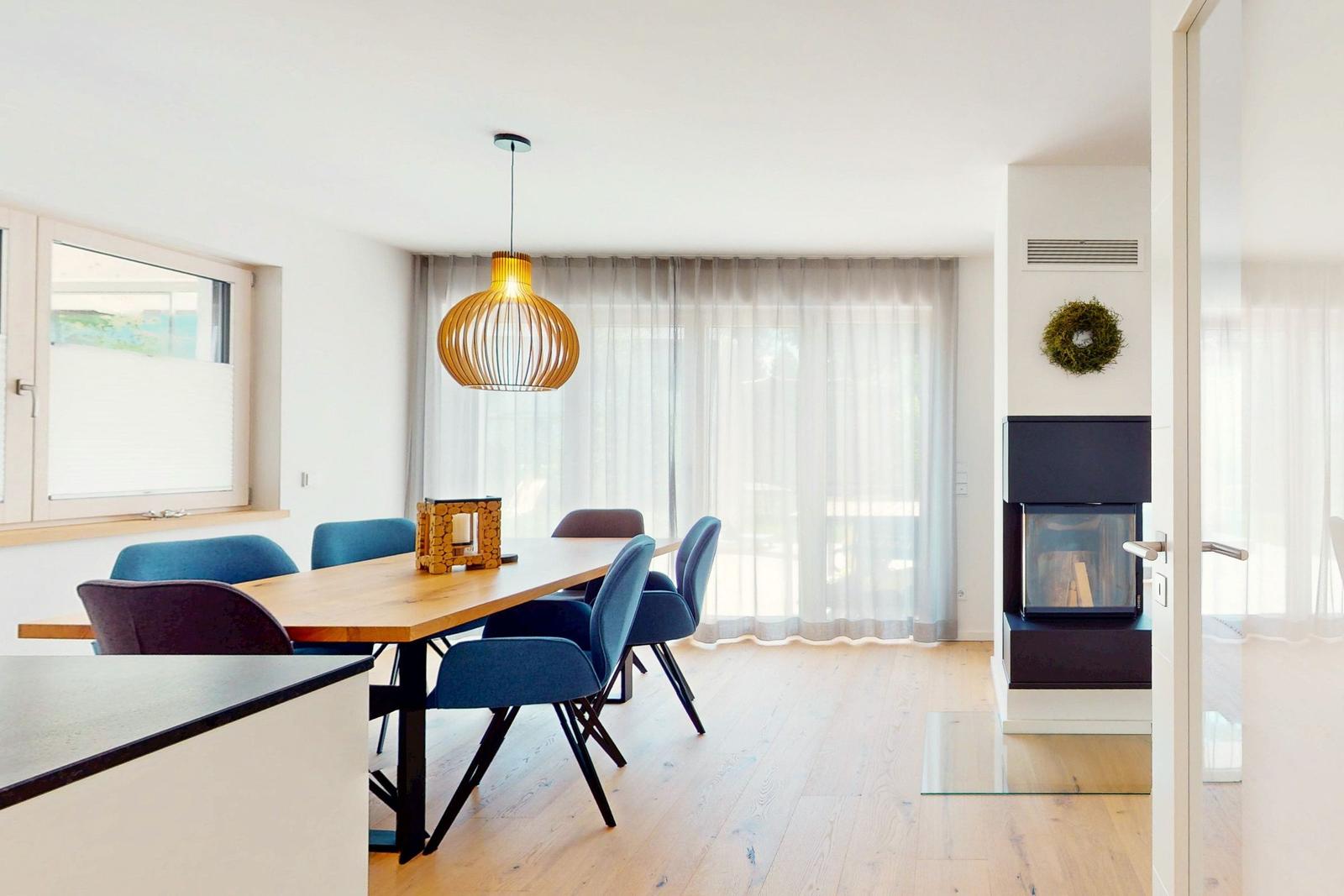 Esszimmer mit Holztisch, blauen Stühlen und Deckenleuchte. Im Hintergrund steht ein Kamin.