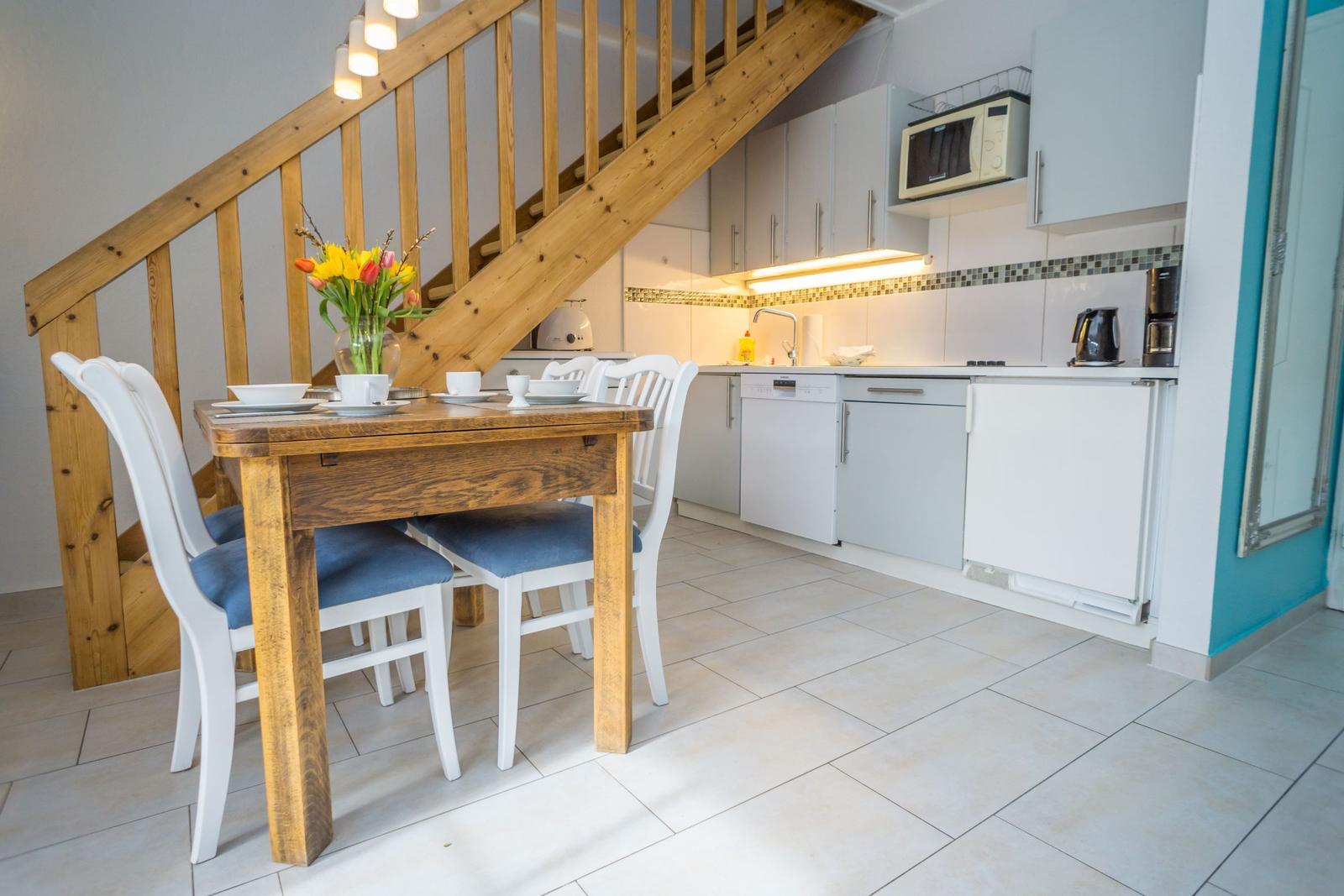 Essbereich mit Holztisch und Stühlen unter einer Treppe. Küche mit weißen Schränken und Küchengeräten.