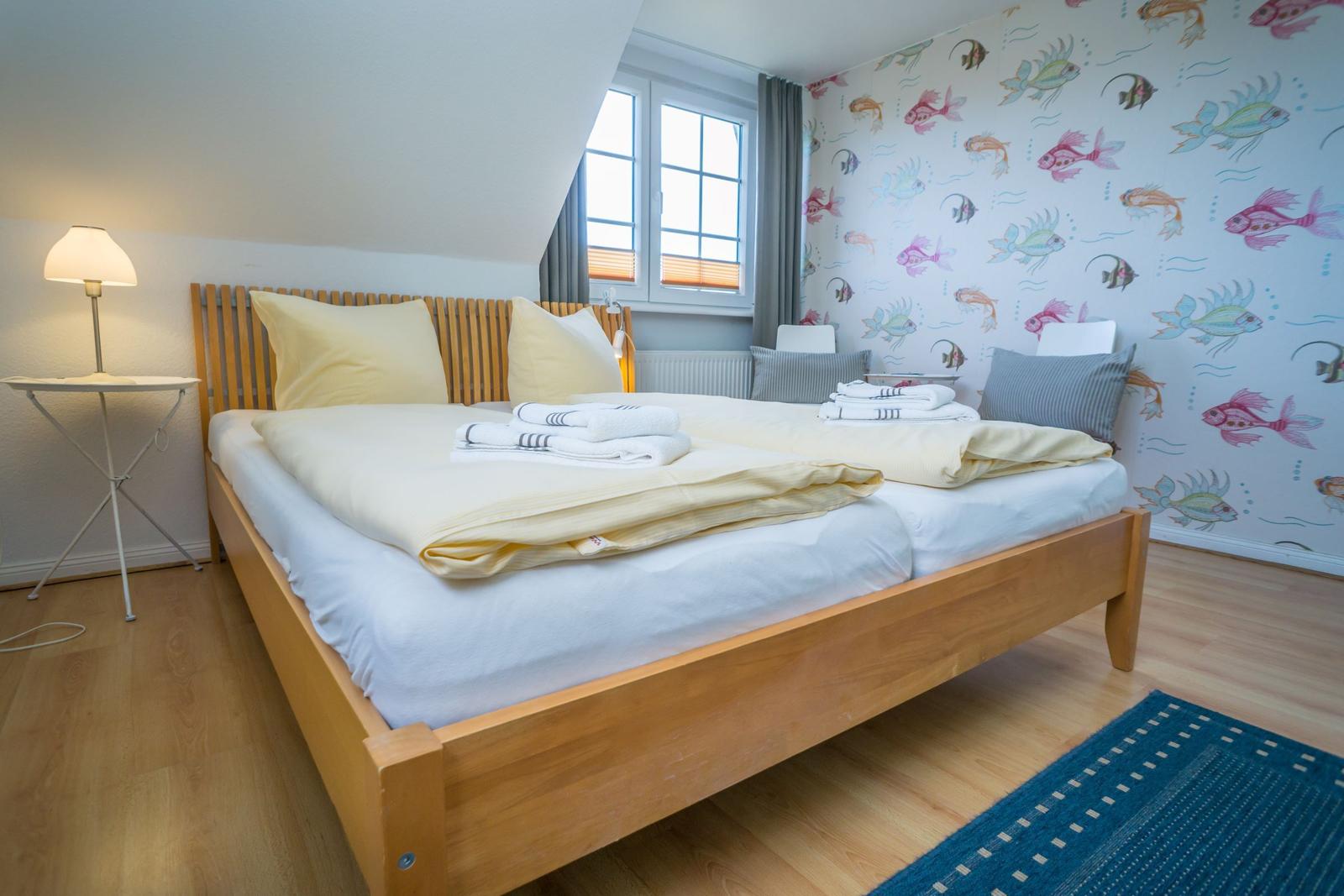 Schlafzimmer mit Holzbett, Fischmotiv-Wand und Fenster mit Jalousie.