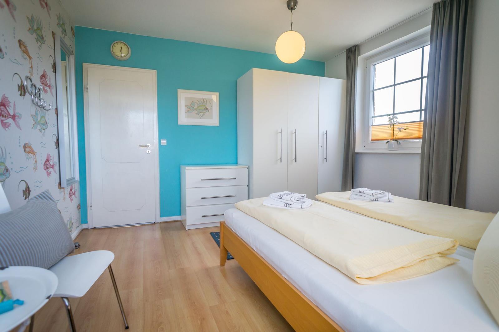 Schlafzimmer mit zwei Betten, blauer Wand, Kleiderschrank und Fenster mit Vorhängen.