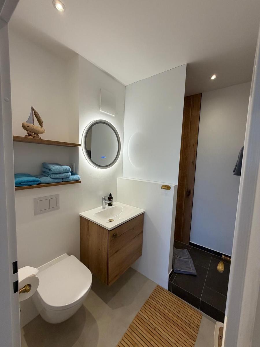 Badezimmer mit Waschbecken, Toilette und Dusche. Holzregal, runder Spiegel und Badezimmermatte.