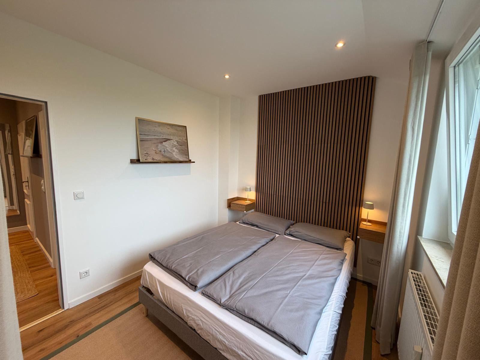 Schlafzimmer mit Doppelbett, Holzpaneelen und Fenster. Links Tür zur angrenzenden Räumlichkeit.