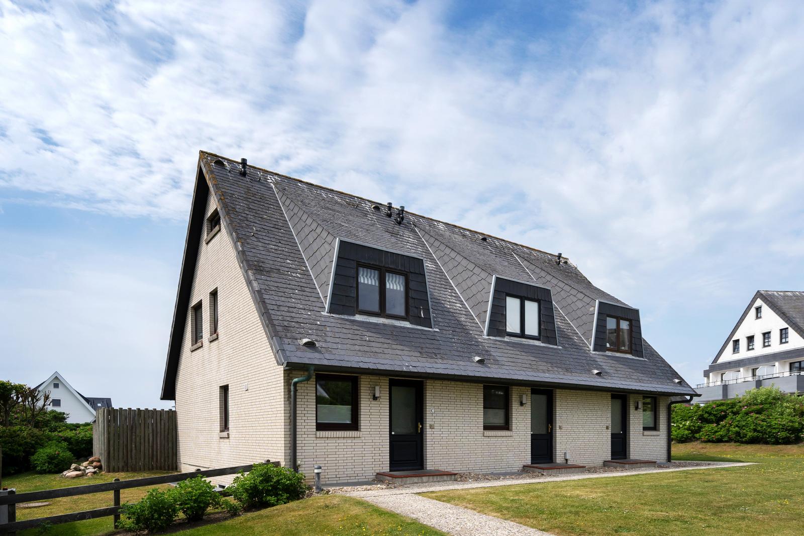 Ein Haus mit schwarzem Dach und hellen Wänden, mehreren Fenstern und einer grünen Umgebung.