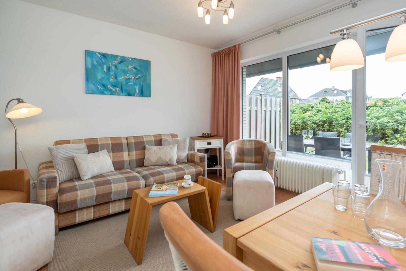 Wohnzimmer mit Sofa, Sessel und Tisch. Große Fensterfront mit Blick auf Garten.