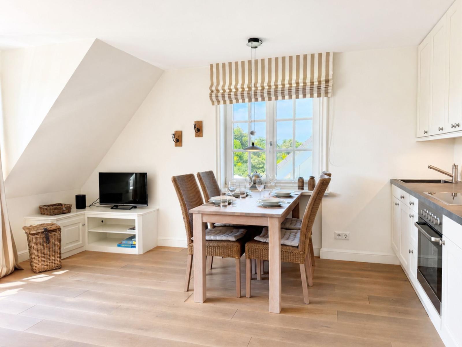 Esszimmer mit Tisch, Stühlen und Fenster. Küche mit Arbeitsplatte und Herd. TV und Korb im Raum.