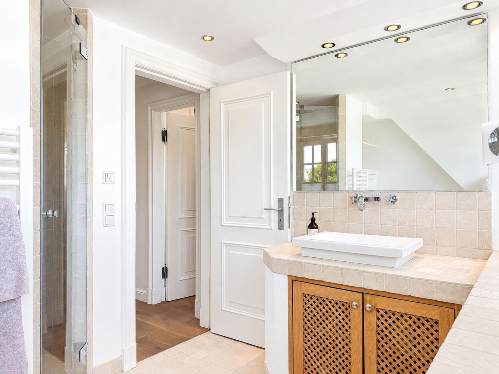Badezimmer mit Dusche, Waschtisch und Spiegel. Holzschrank und hellen Fliesen.