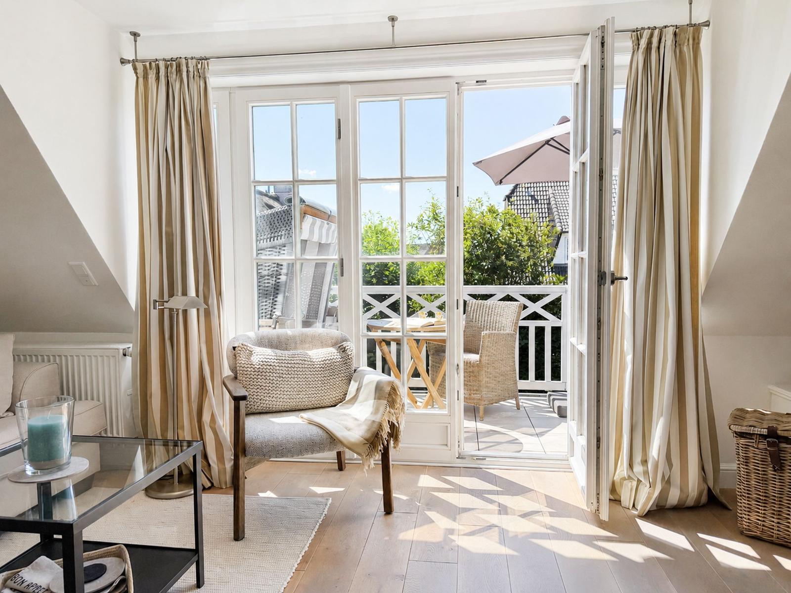 Ein Raum mit großem Fenster und Balkon. Vorhänge, Sessel und Tisch sind sichtbar.