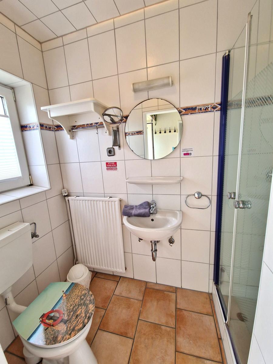 Badezimmer mit Dusche, Waschbecken und Toilette. Fliesenwand und Fenster sichtbar.