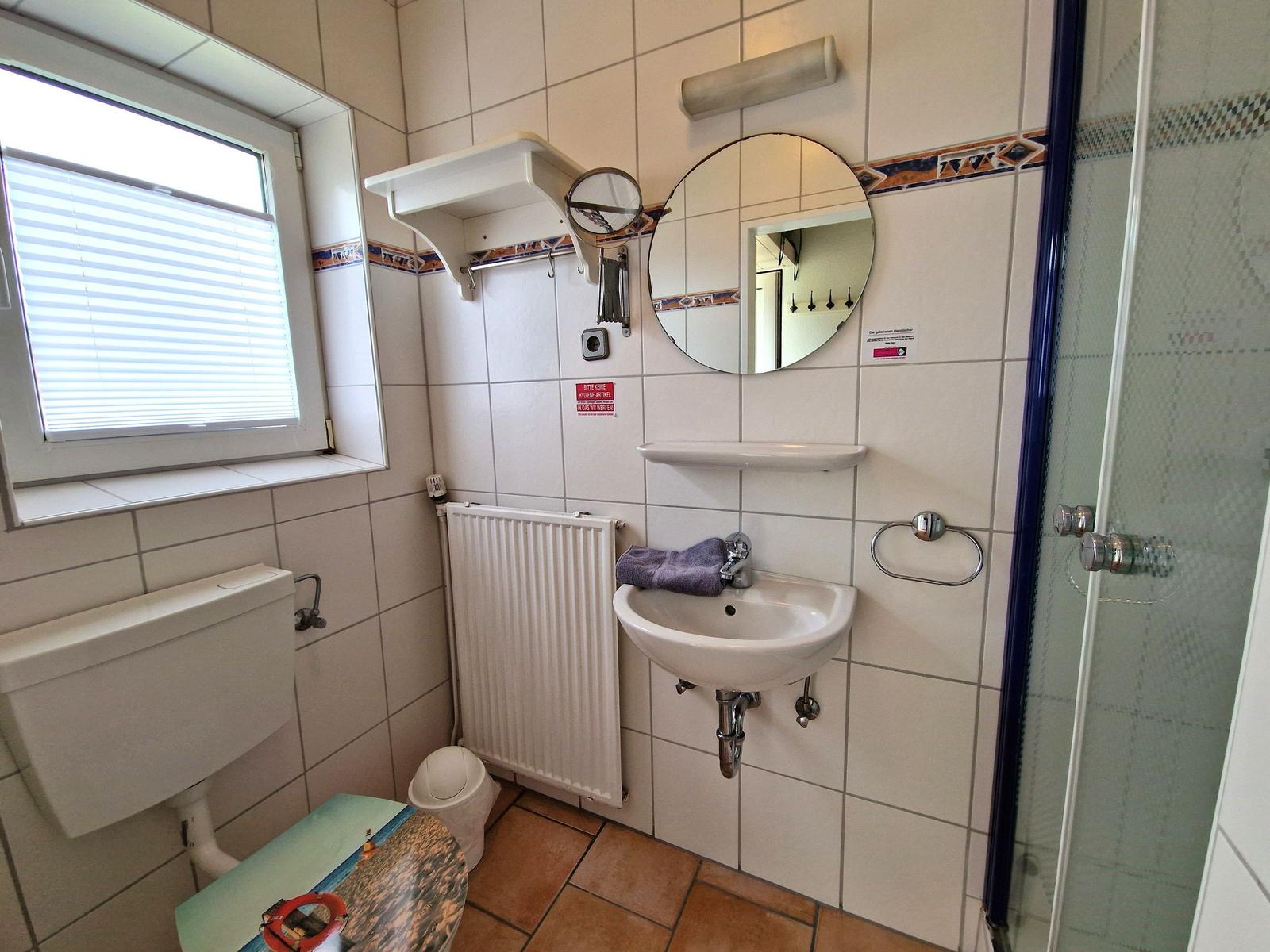 Badezimmer mit Dusche, Waschbecken und Toilette. Fenster mit Jalousie.