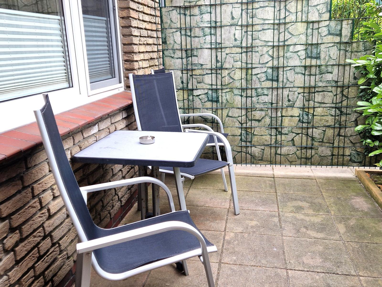 Terrasse mit Tisch und zwei Stühlen, Steinmauer und Pflanzen im Hintergrund.