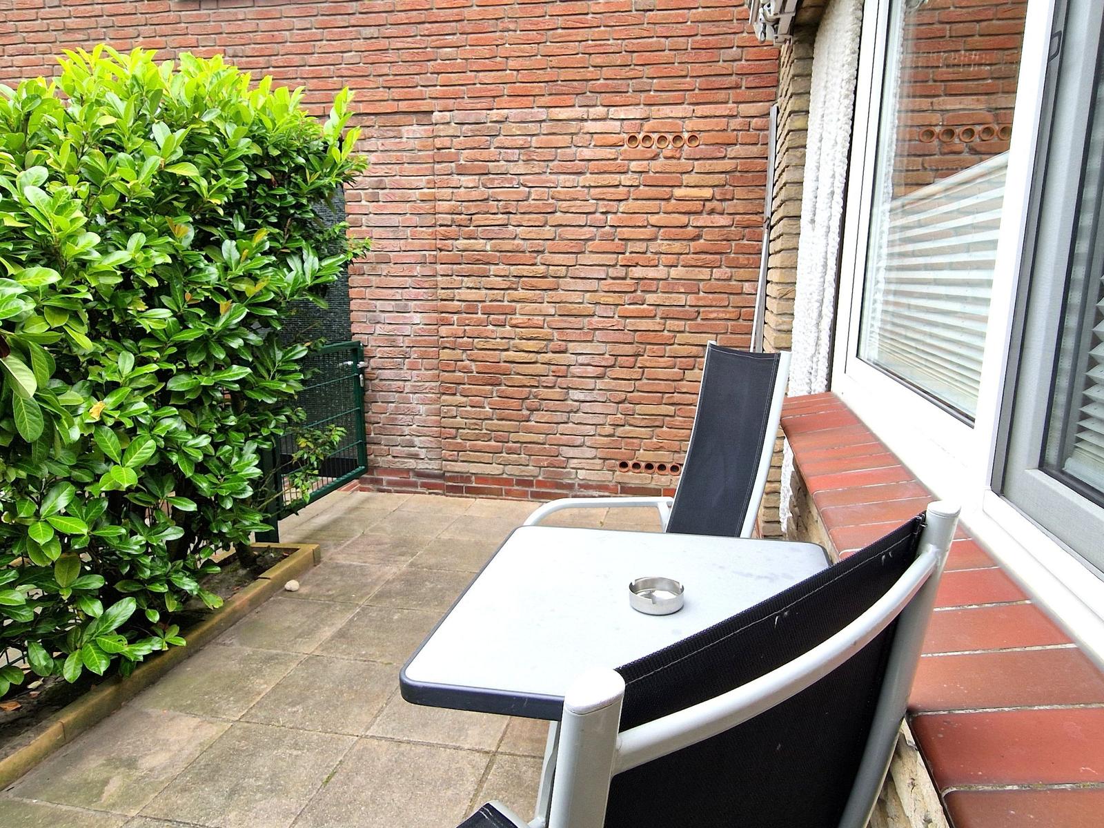 Terrasse mit Tisch, zwei Stühlen und Aschenbecher. Hintergrund: Ziegelwand und grüner Strauch.