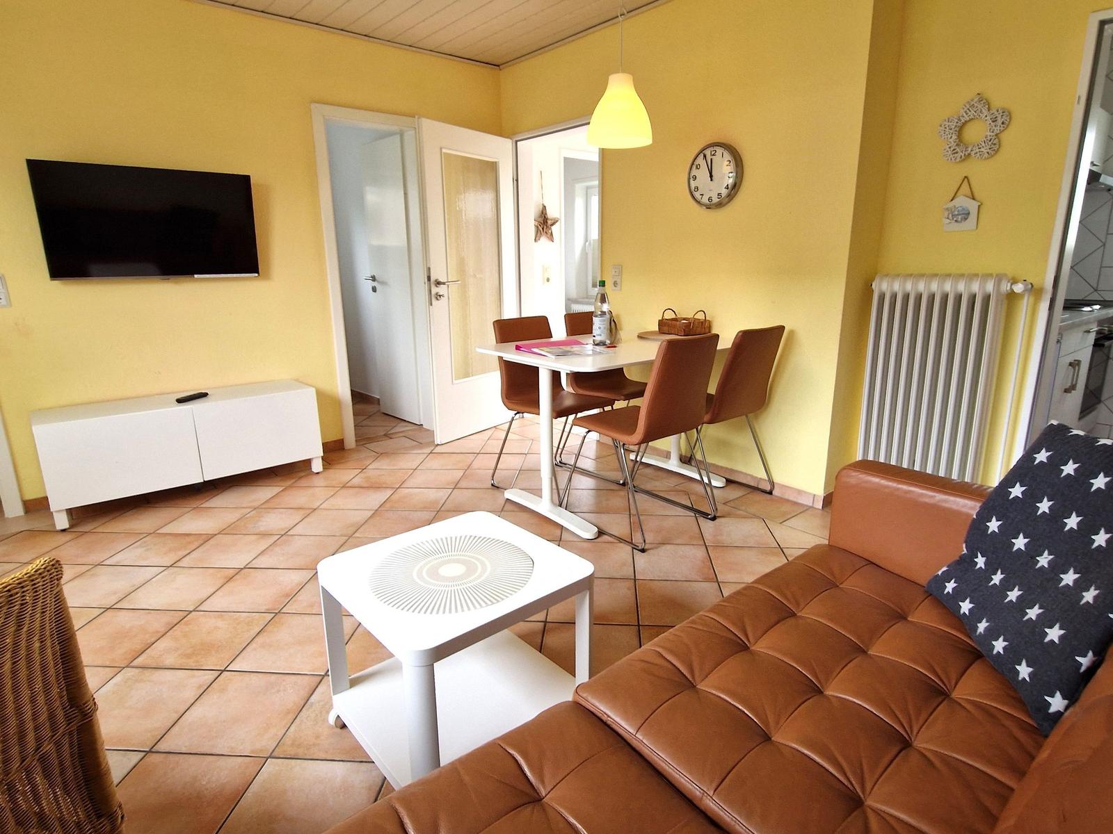 Wohnbereich mit Sofa, Esstisch und Fernseher an gelber Wand.