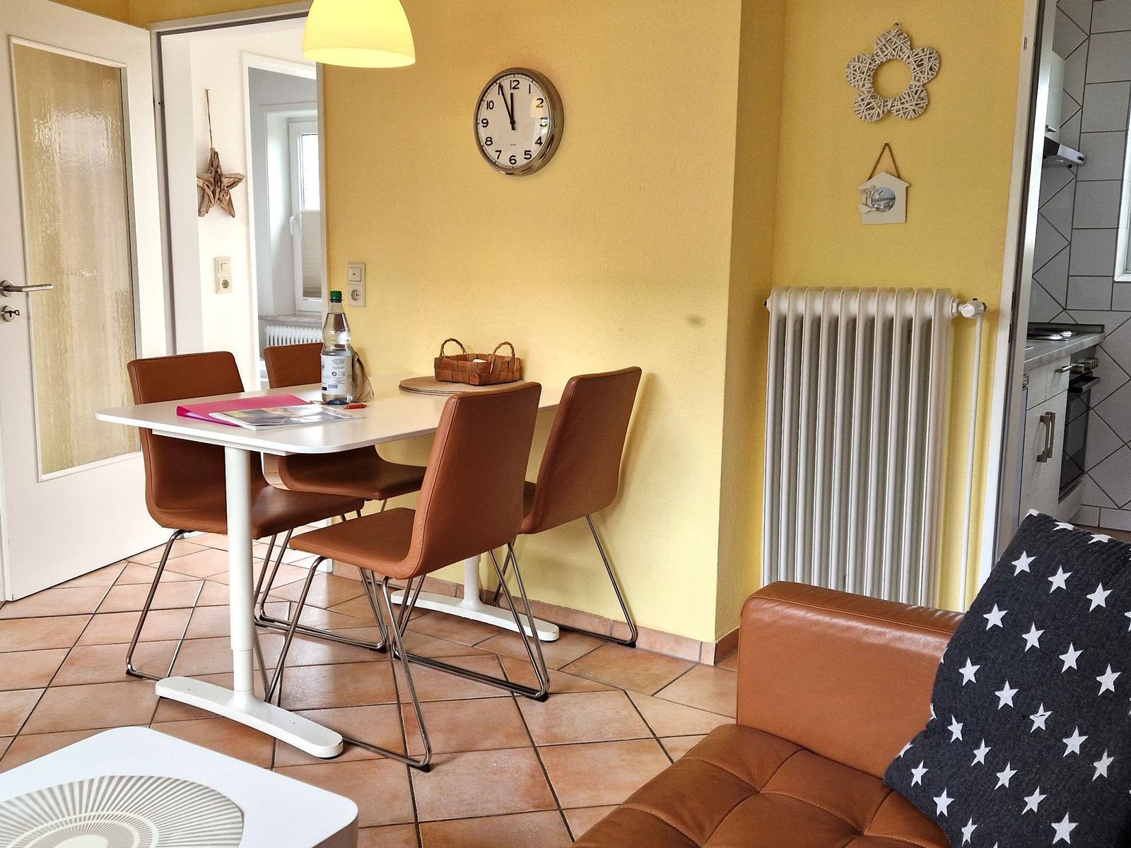 Essbereich mit Tisch, vier Stühlen und Wanduhr. Kaminofen und Sofa im Vordergrund.