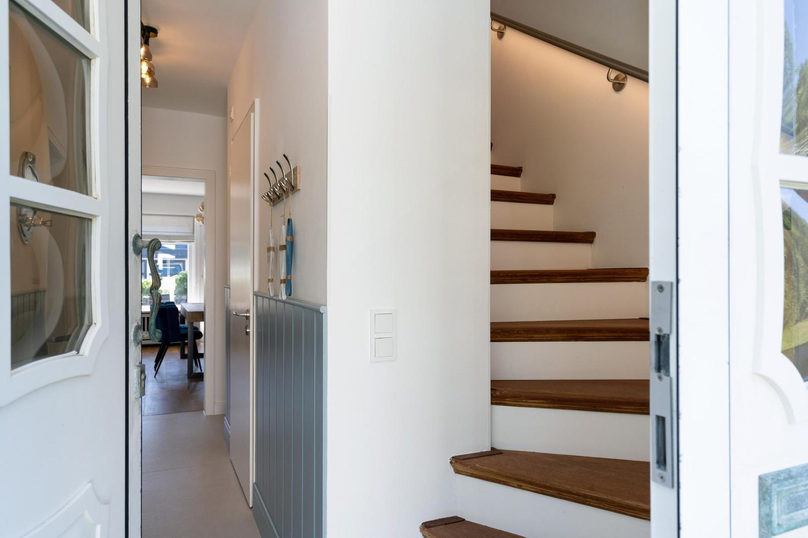 Hausgang mit Holztreppe und weißen Wänden. An der Wand hängen Haken mit blauen Gegenständen.