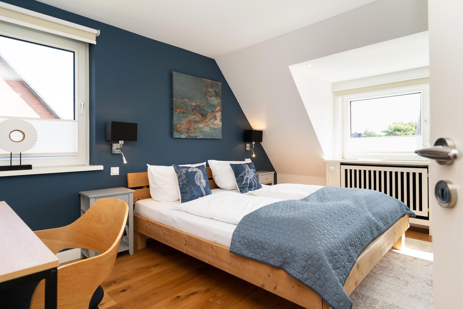 Schlafzimmer mit Holzbett, blauer Wand und zwei Fenstern.