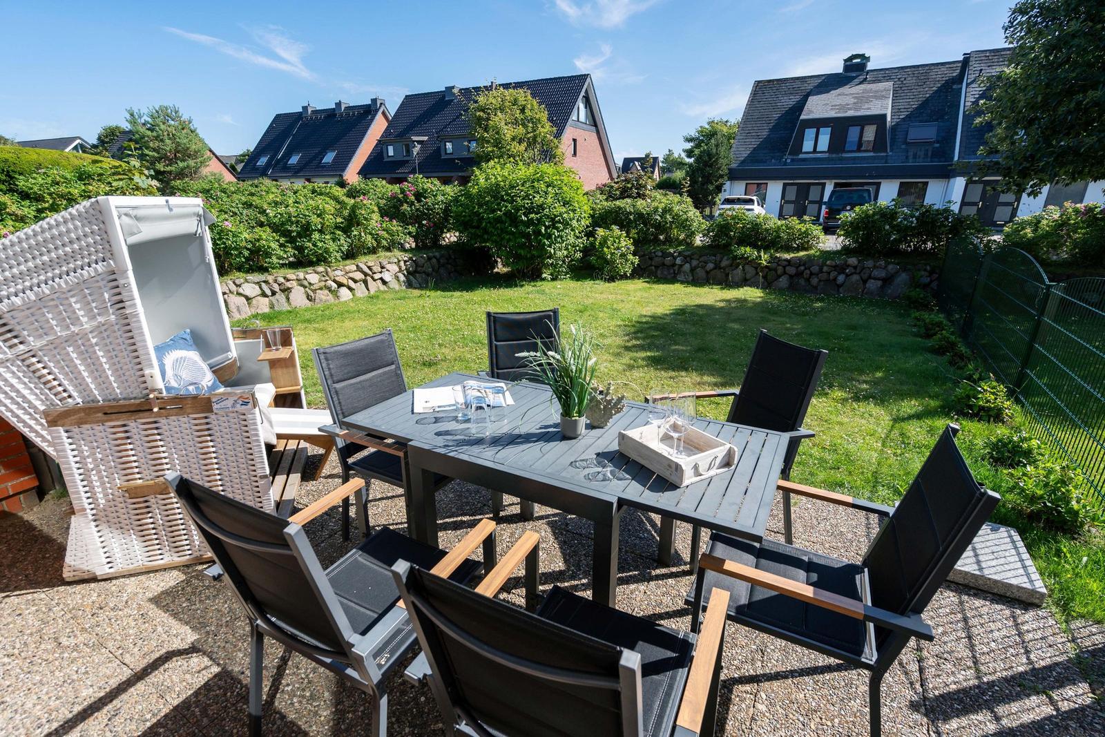 Terrasse mit Tisch, Stühlen und Strandkorb. Grünfläche und Häuser im Hintergrund.