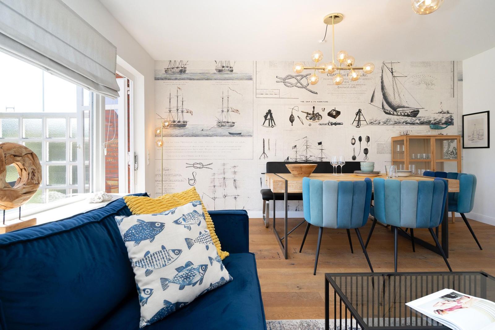 Wohn- und Essbereich mit blauem Sofa, Holztisch und blauen Stühlen. Wand mit Schiffsmotiven.