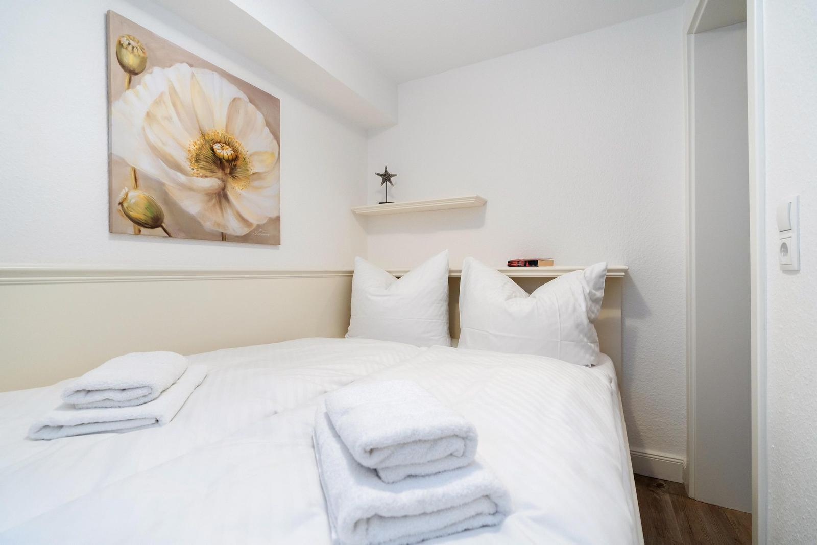 Schlafzimmer mit weißem Bett, Kissen und Handtüchern. Wandbild mit Blume und kleine Ablage.
