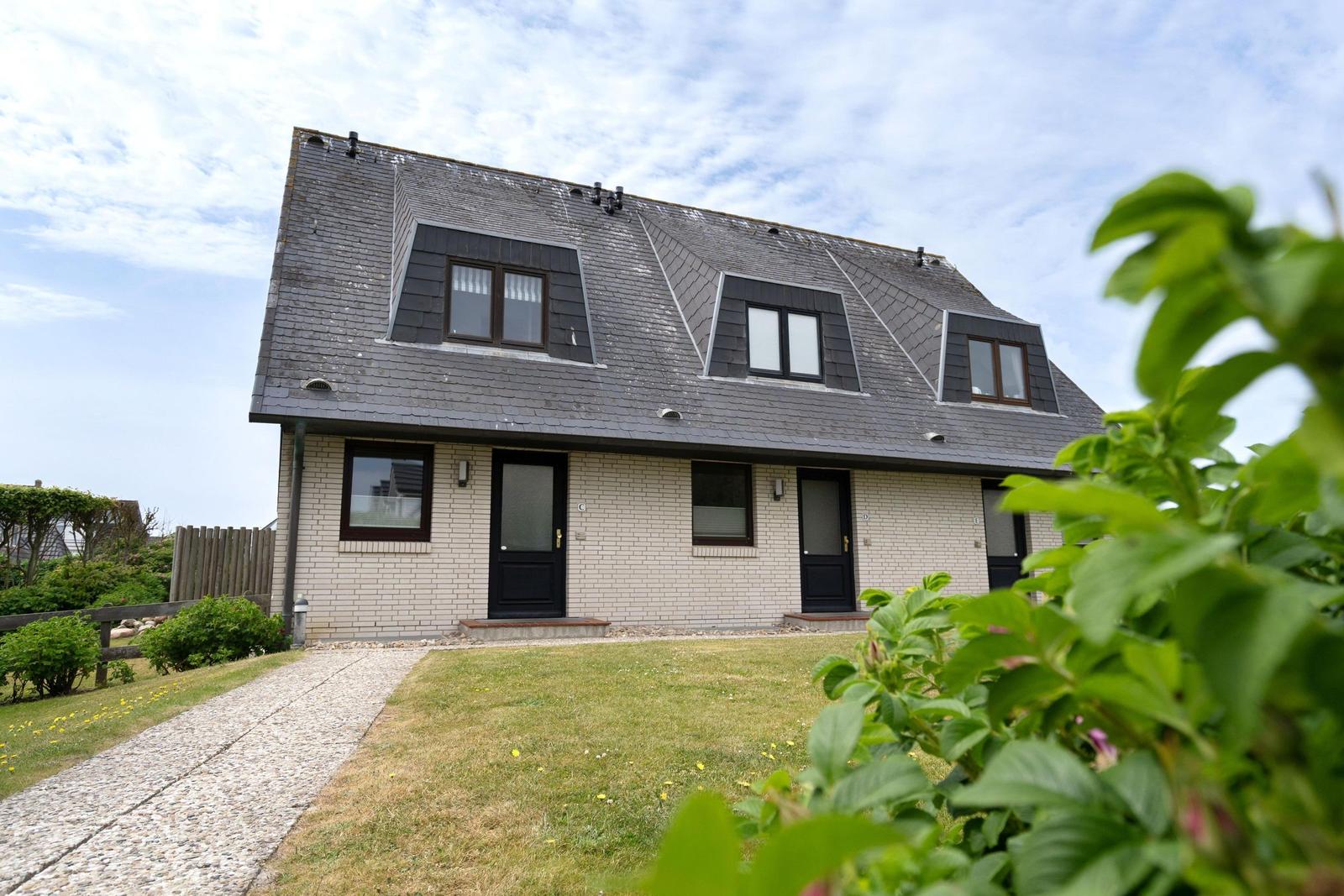 Ein Haus mit grauem Dach und hellen Wänden, umgeben von Grün und einem Kiesweg.