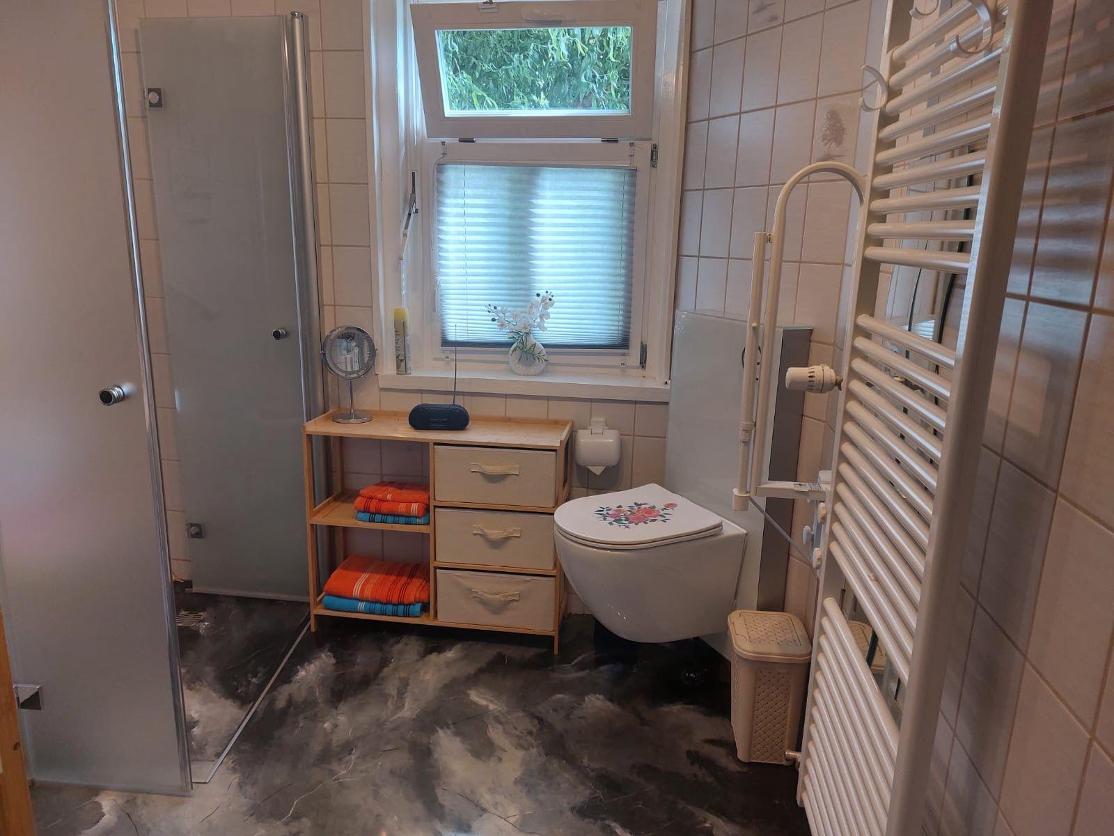 Badezimmer mit Dusche, Toilette und Heizkörper. Fenster mit Jalousie und Handtuchregal.