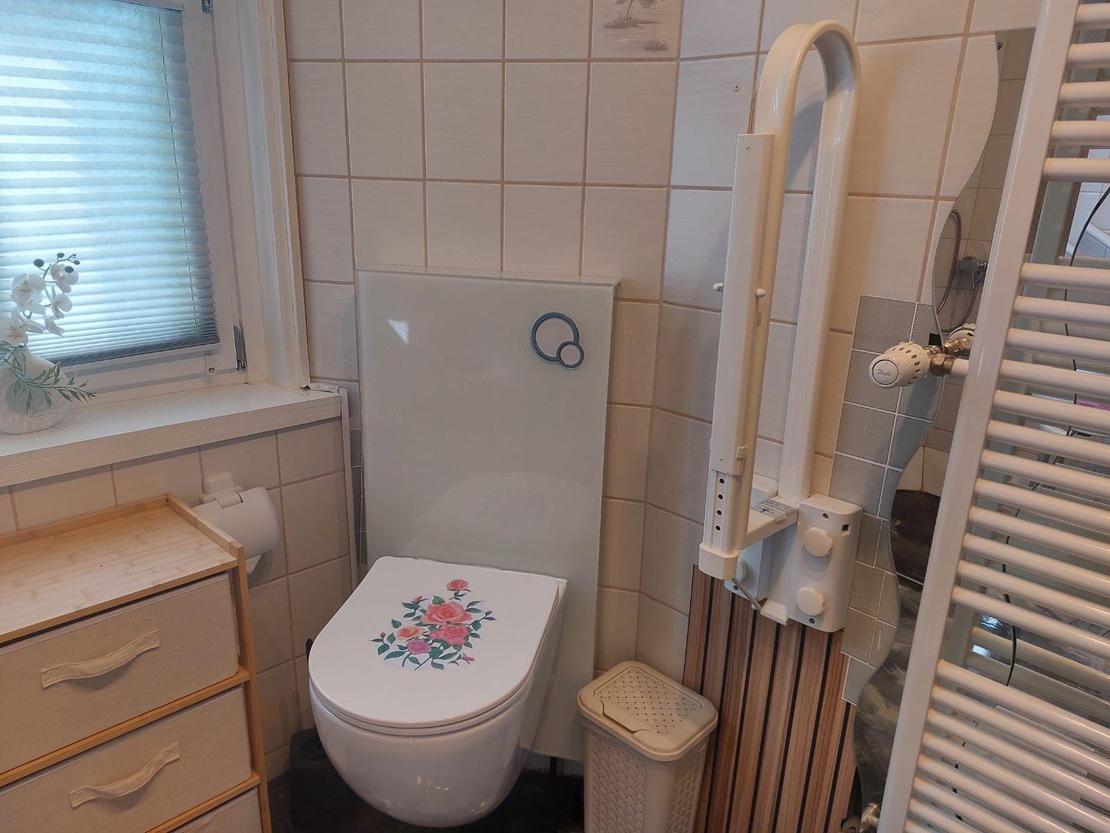 Bad mit Toilette, Duschbereich und Heizung. Fenster mit Jalousie und Blumen.