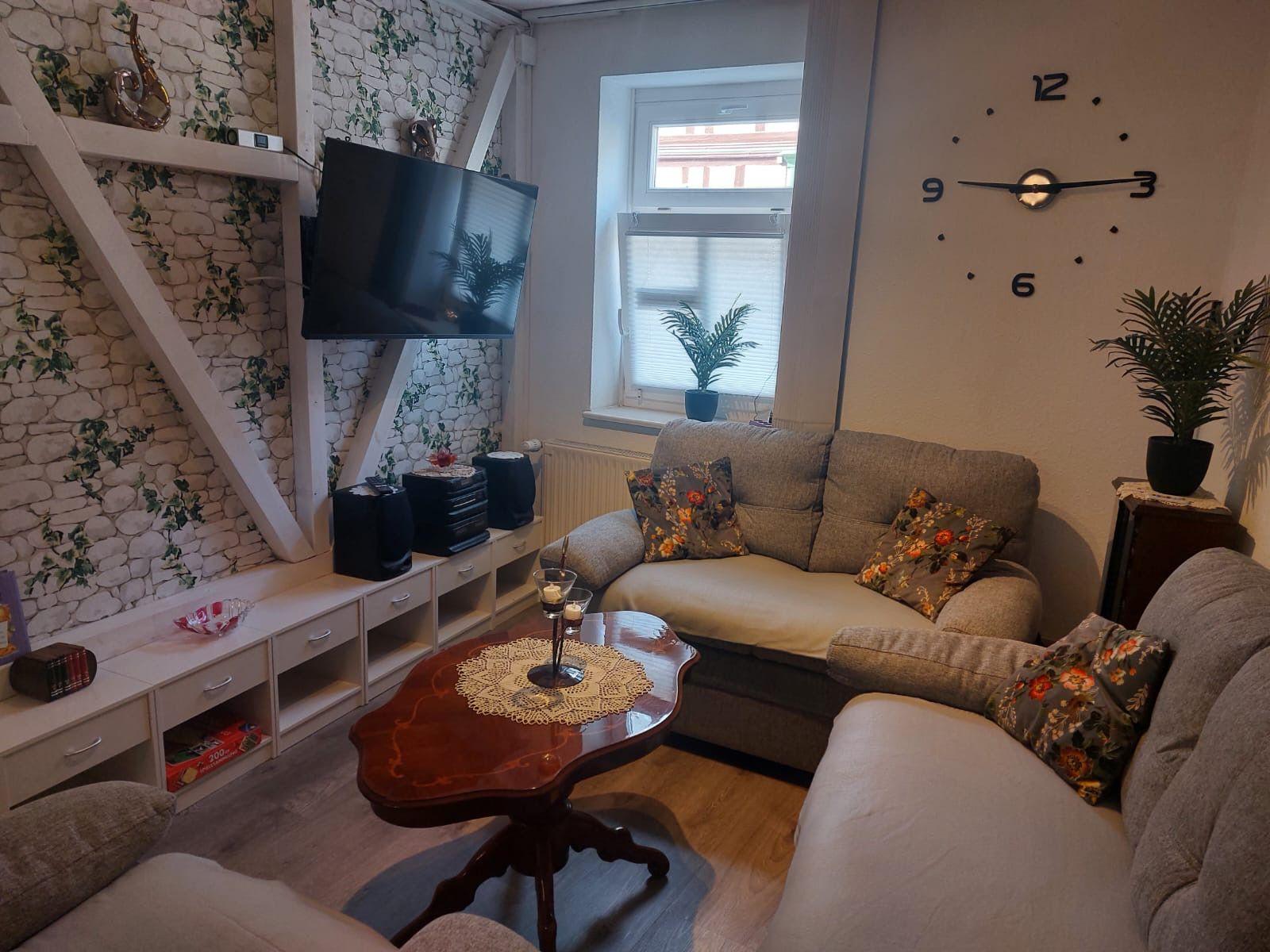 Wohnzimmer mit Sofa, Fernseher und Tisch. Wanduhr und Pflanzen sichtbar.