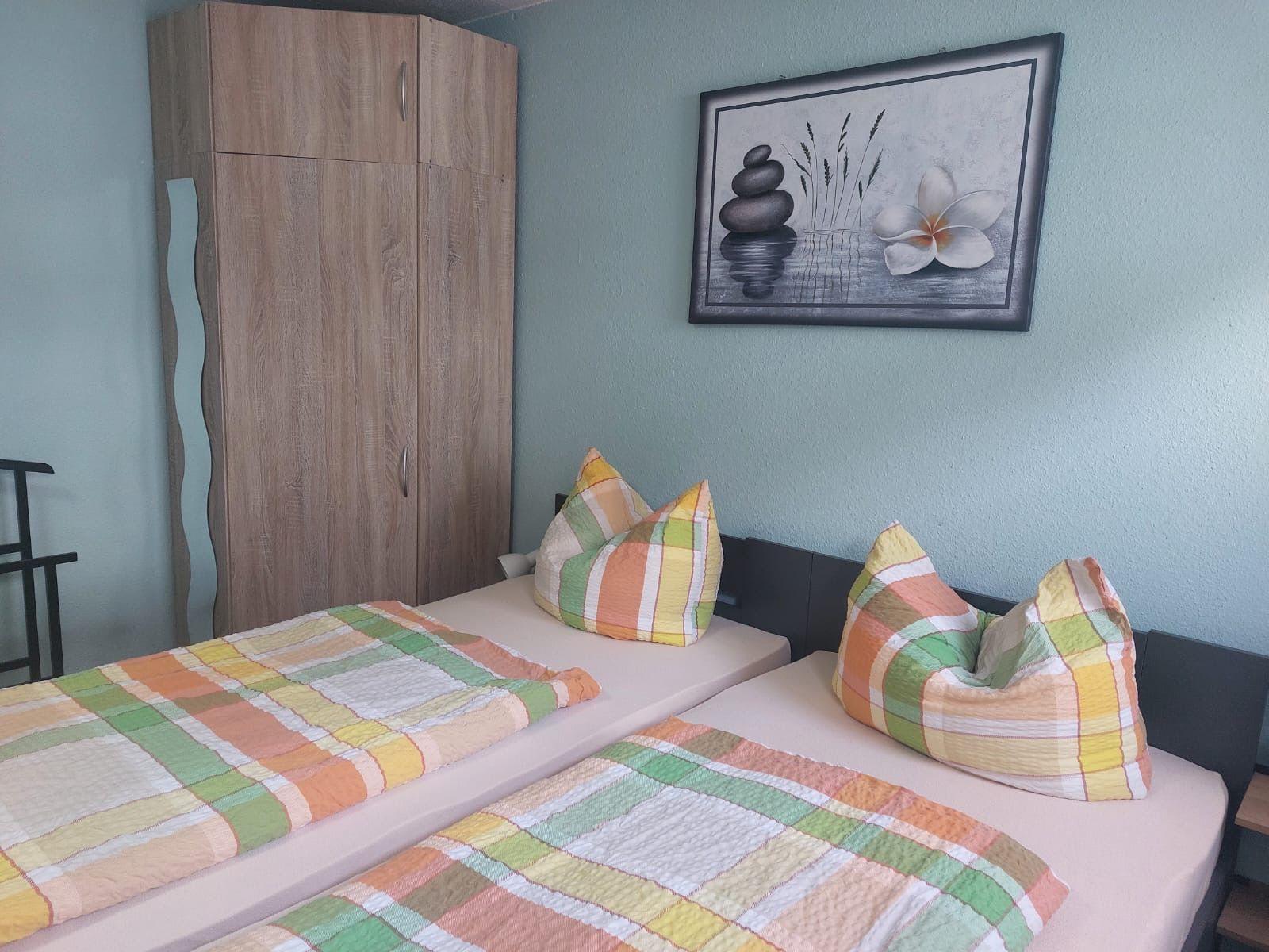 Zwei Betten mit bunten Bettwäschen, Kleiderschrank und Wandbild mit Stein- und Blumenmotiv.