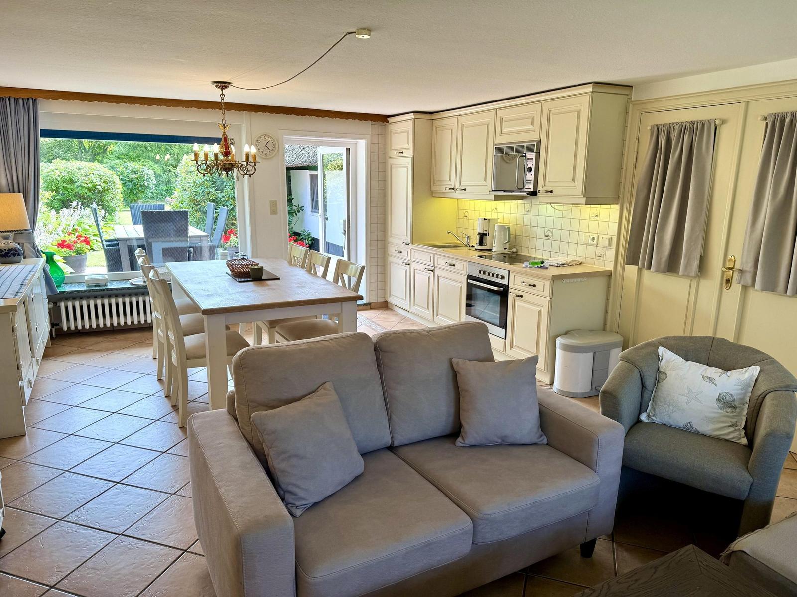 Living and dining area with kitchen, sofa, and armchair. Passage to terrace with garden view.