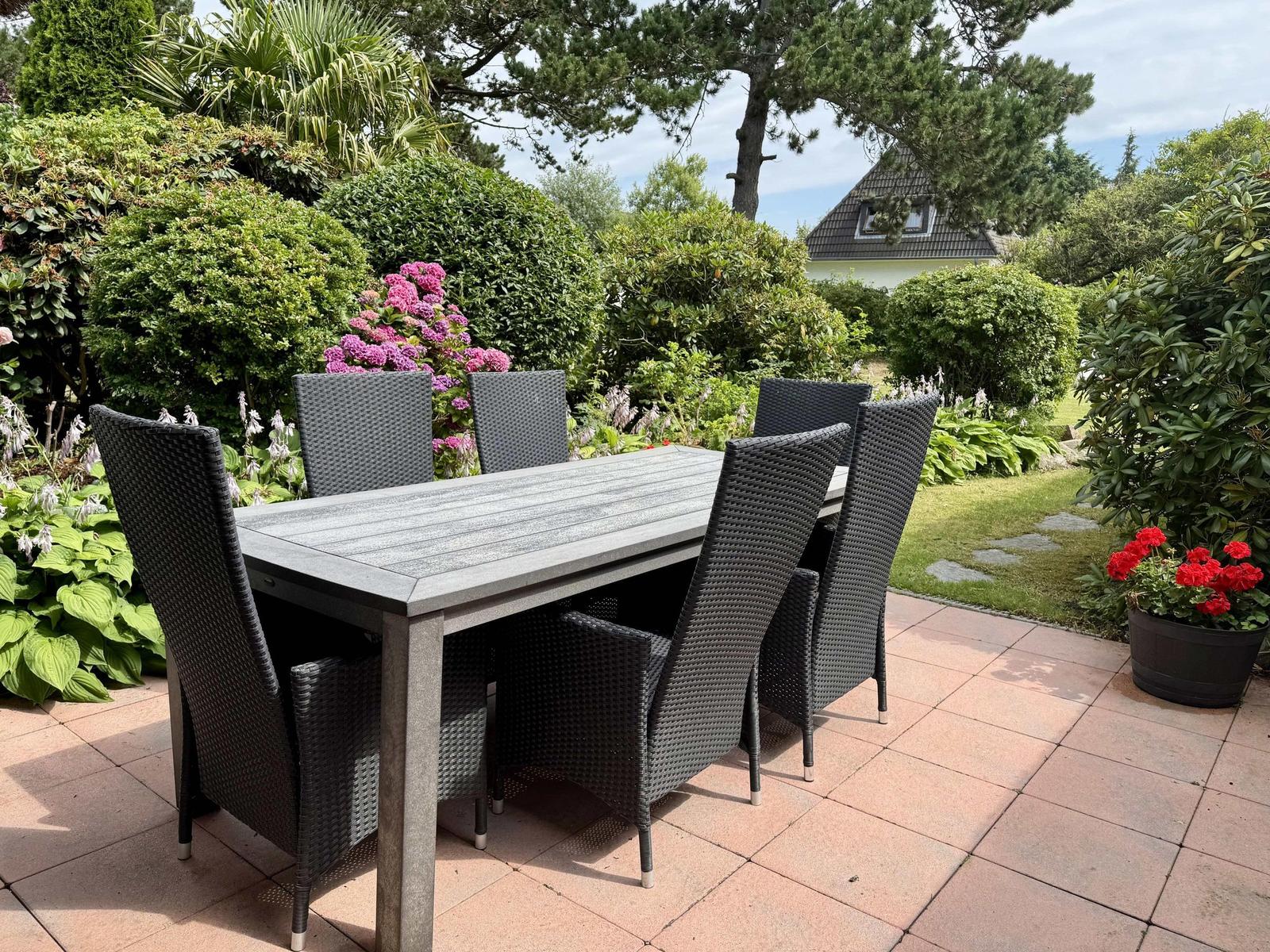 Outdoor area with covered table and six chairs. Surrounded by flowers and greenery.