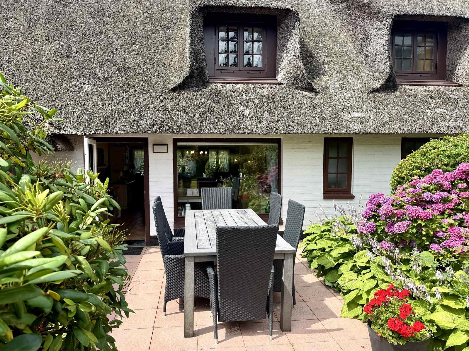 Terrace with table and chairs in front of a house with thatched roof and flower beds.