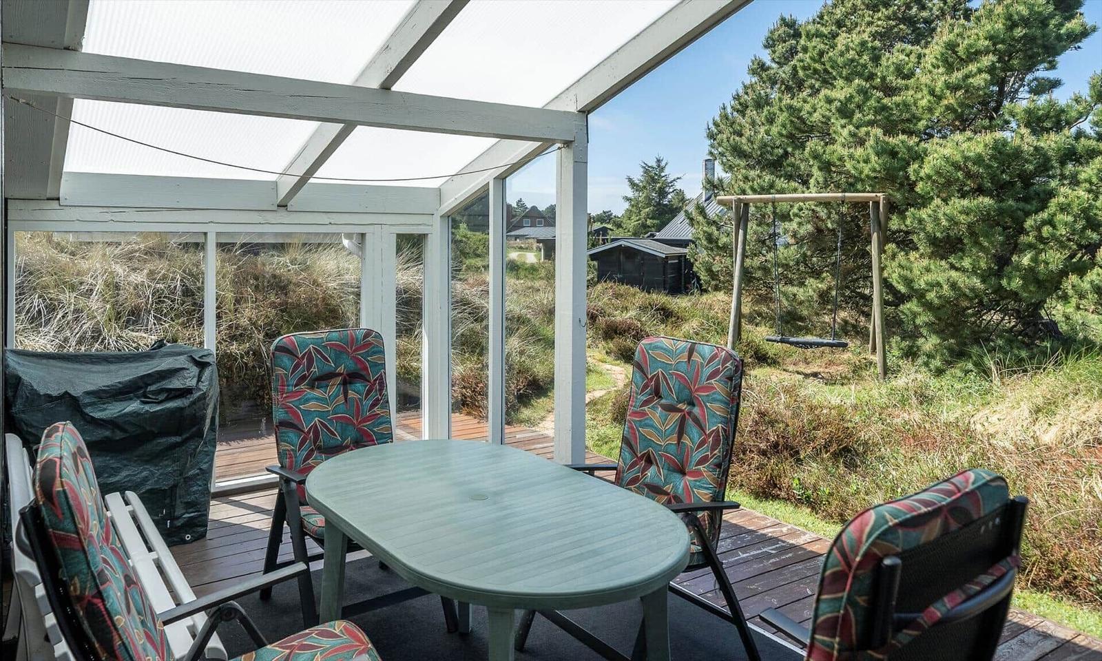 Terrasse mit Tisch und Stühlen unter Glasdach. Blick auf Grünfläche und Schaukel.