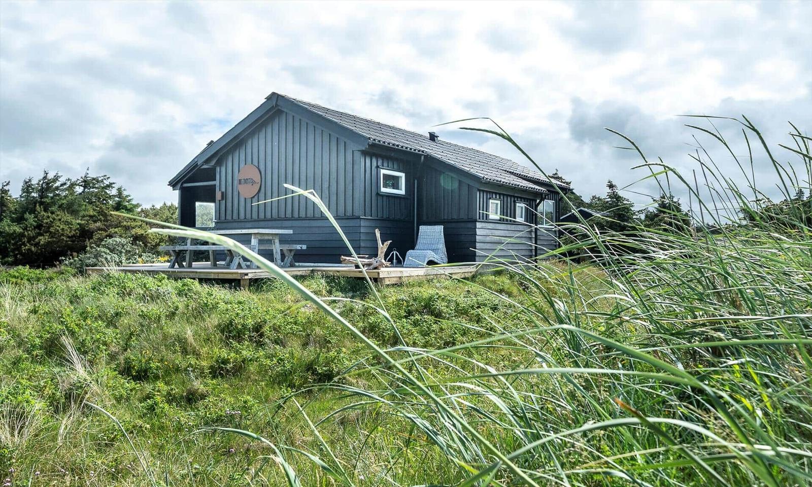 A gray wooden house with a terrace, picnic table, and lounge chair in a green landscape.