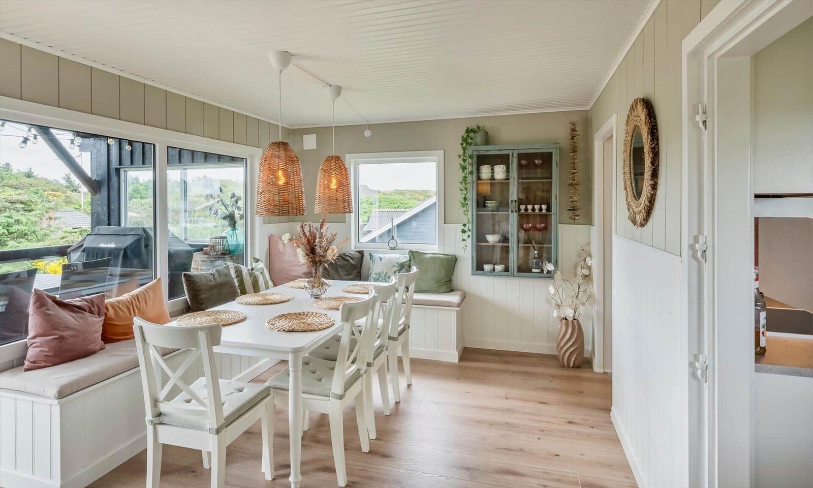 Dining room with white table, chairs, and window seat. Hanging lamps and cabinet are visible.