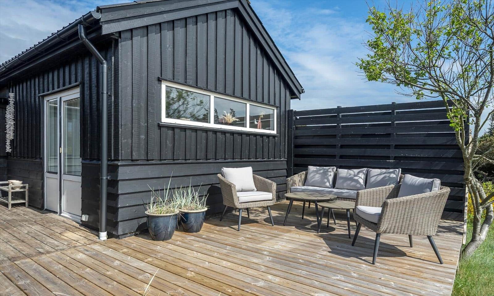 Deck with seating group, wooden floor, and black siding. Plants and tree in background.