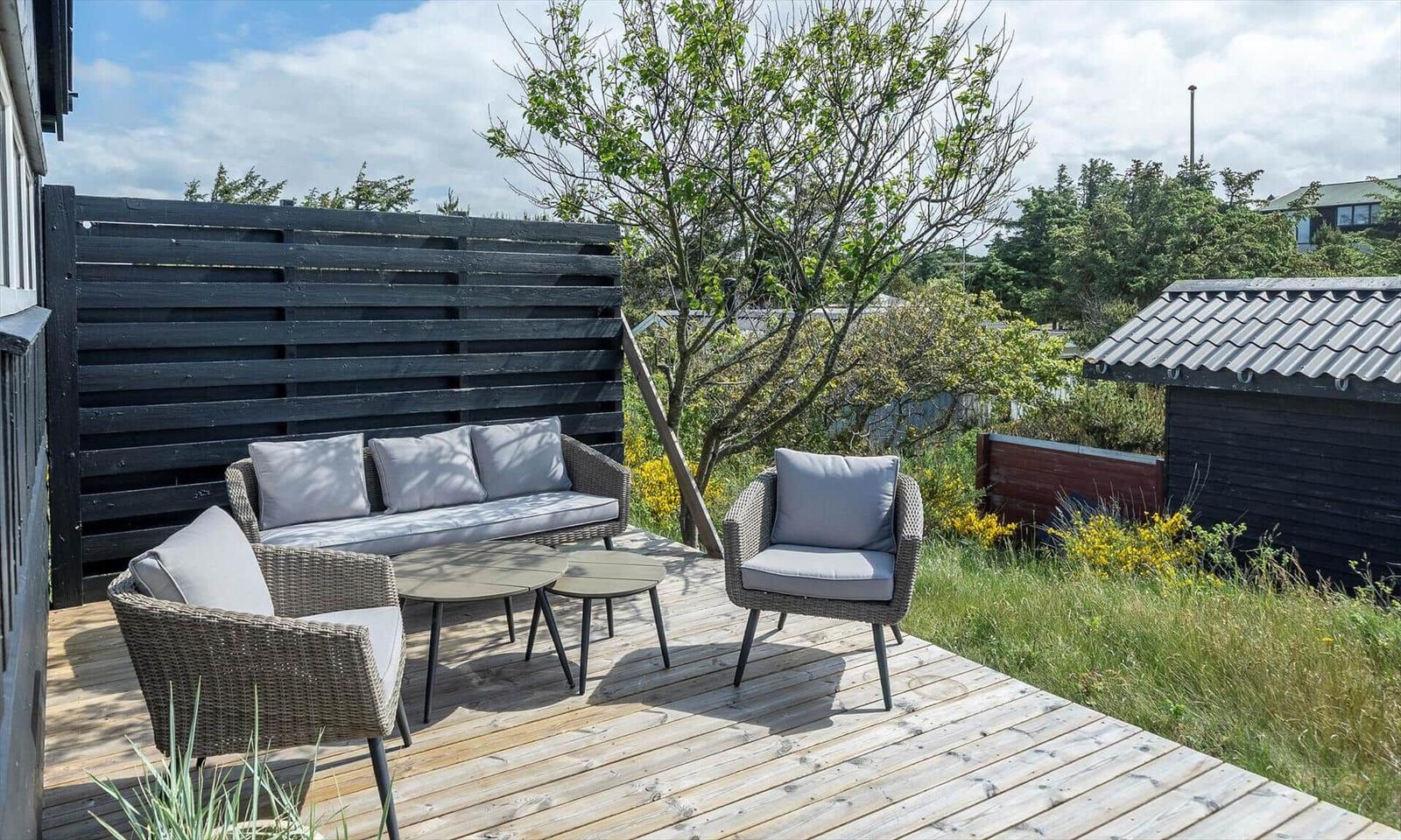 Deck with seating group, table, and wooden railing. Background: greenery and fence.