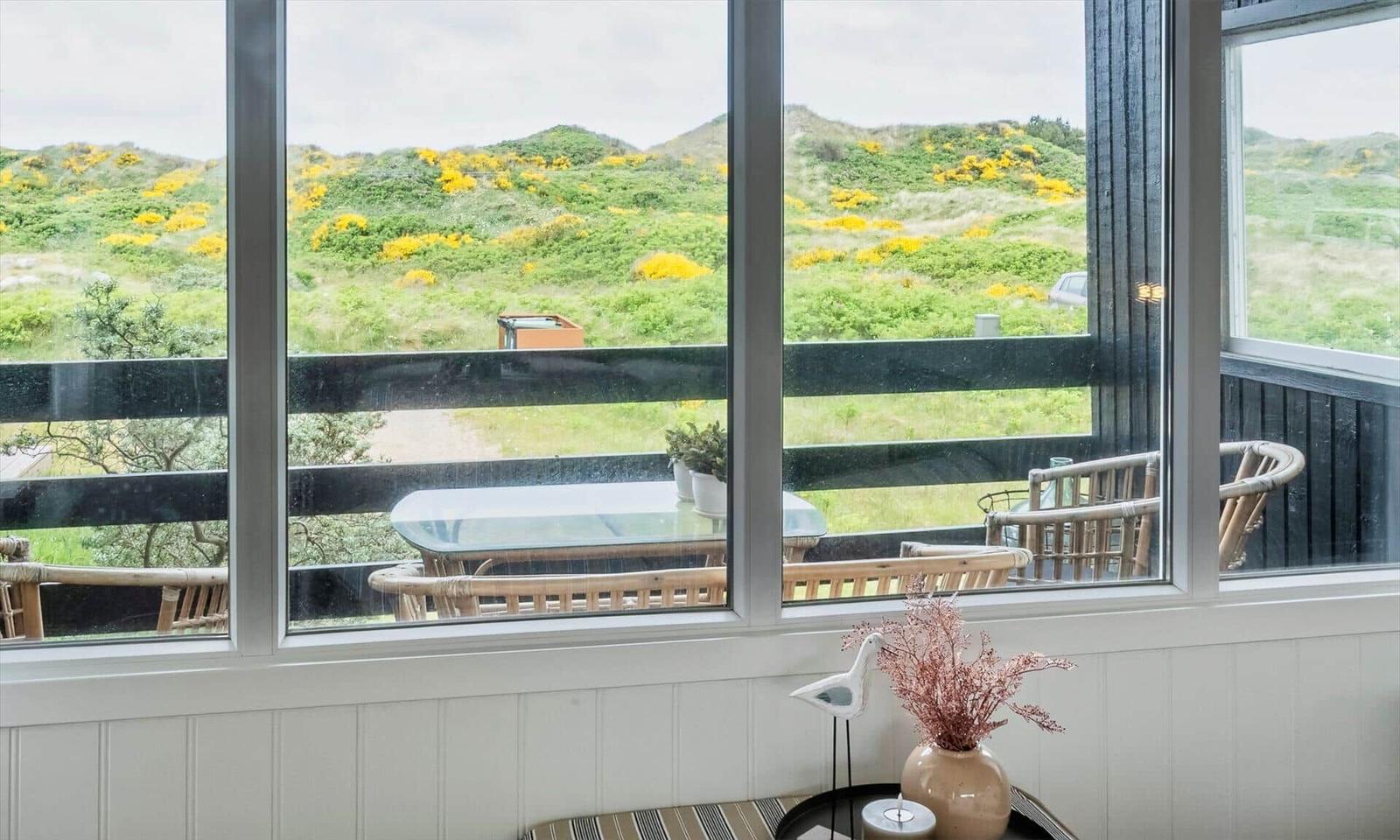Large window with view of green hills and yellow flowers. Balcony with table and chairs.