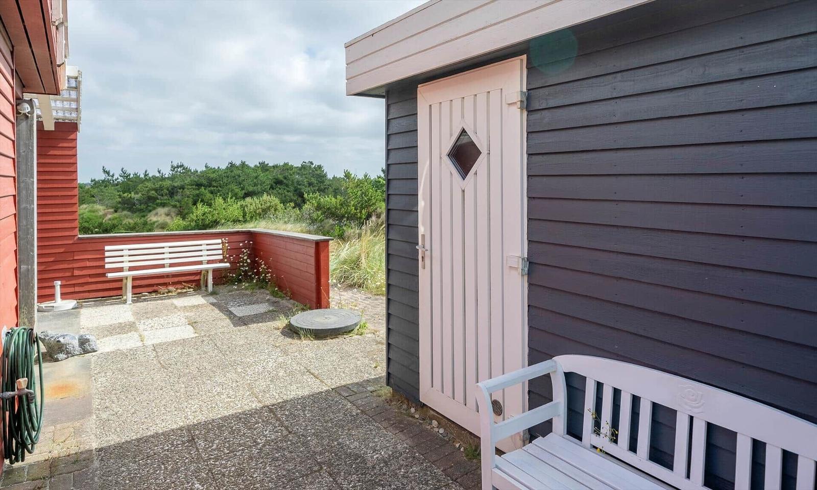 Terrasse mit weißem Bank und grauem Holzhaus. Roter Zaun und grüne Landschaft im Hintergrund.