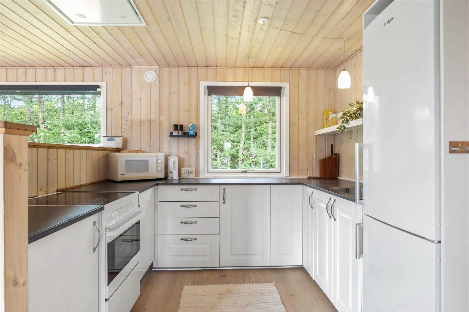 Küche mit weißen Schränken, Holzwand und Fenstern zum Wald.