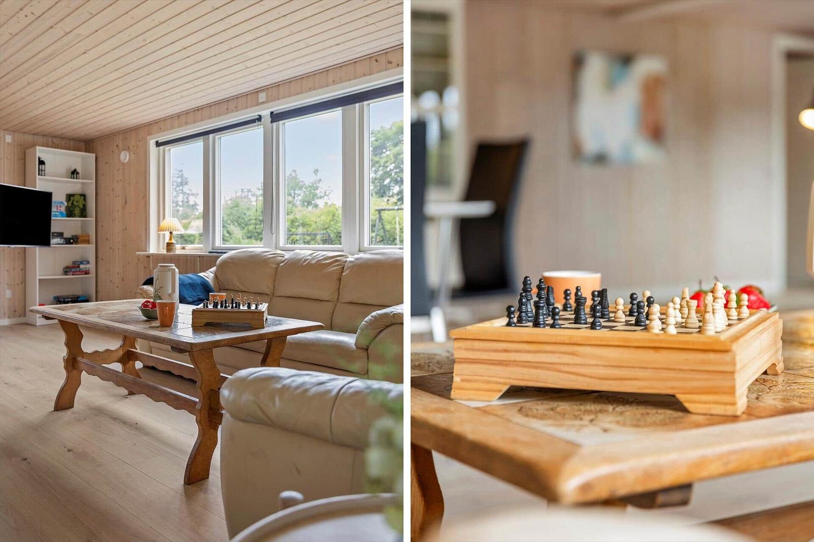 Wohnzimmer mit Holztisch, Sofa und Schachbrett. Große Fenster mit Blick auf Grün.