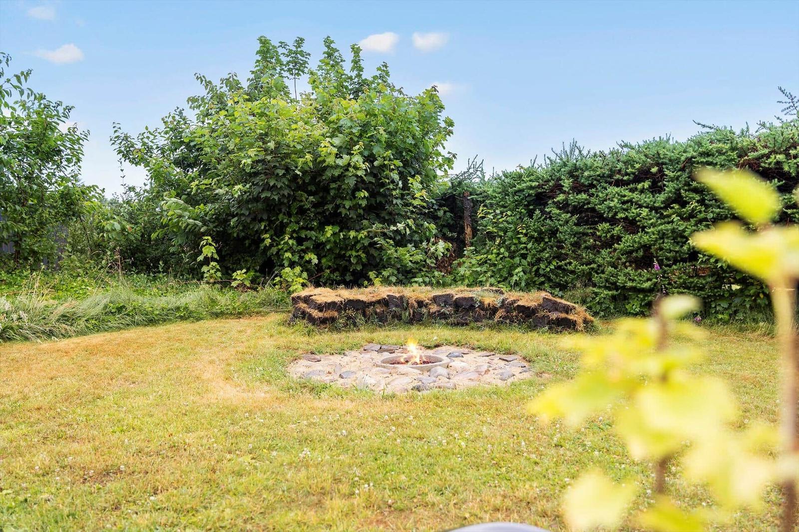 Garten mit Steinsaum und brennendem Feuerkorb umgeben von Gras und Sträuchern.