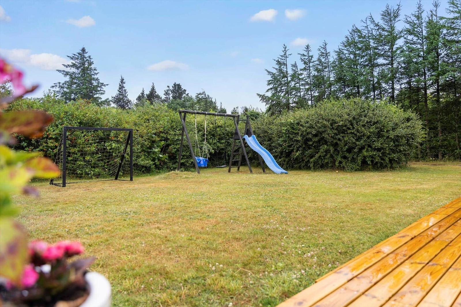 Hinterhof mit Spielplatz, Fußballtor und Holzterrasse. Grünfläche und Bäume im Hintergrund.