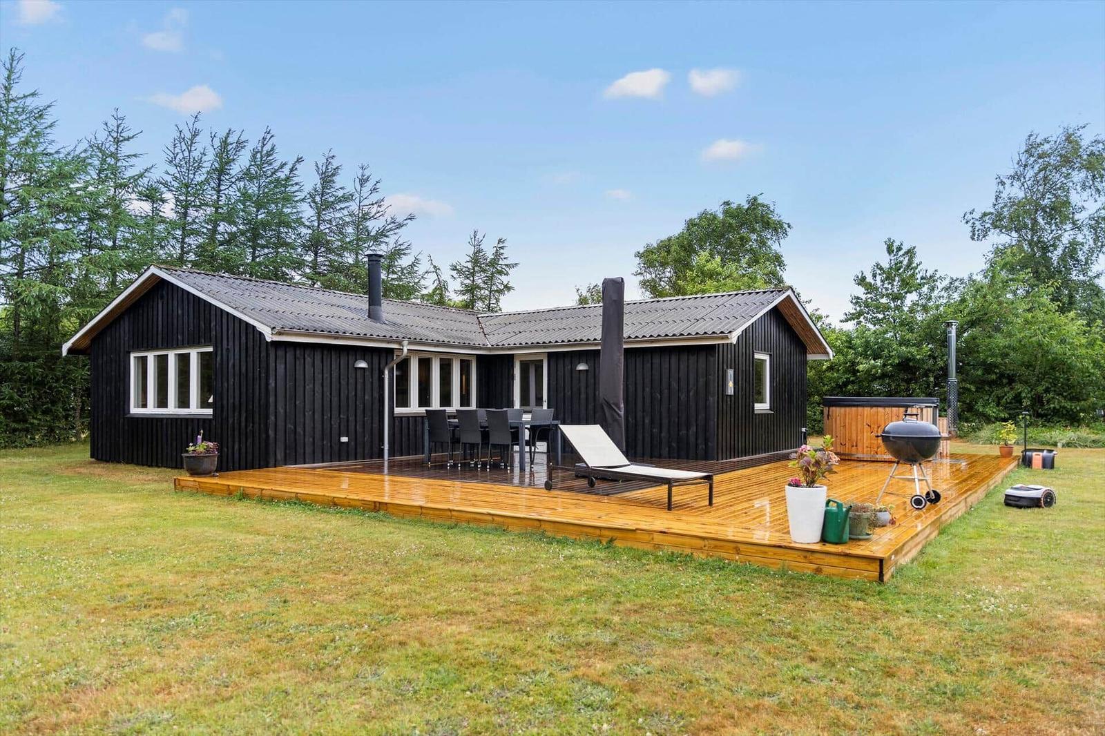 Schwarzes Holzhaus mit Terrasse, Grill und Liege. Umgeben von Gras und Bäumen.