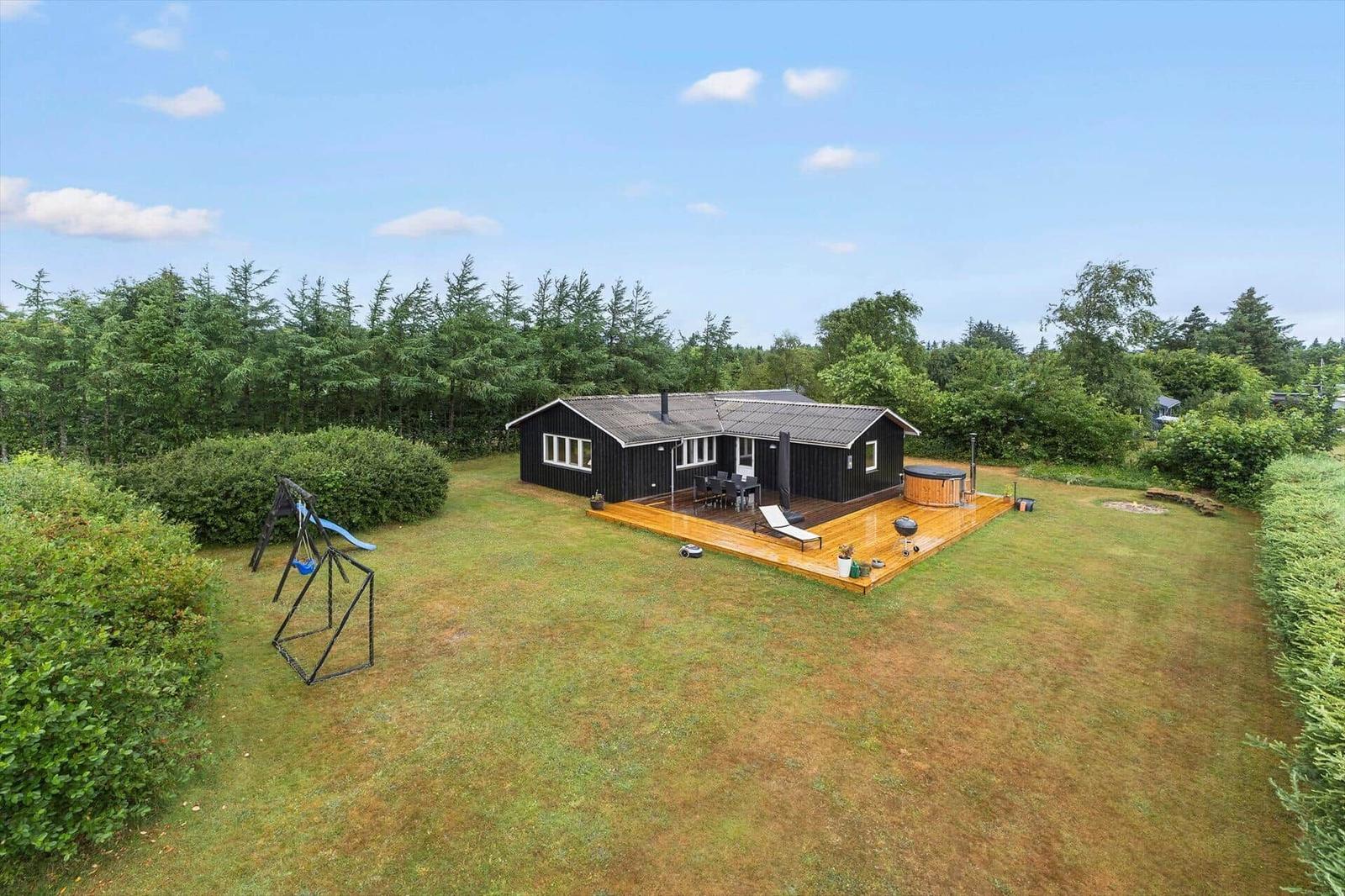 Schwarzes Haus mit Holzterrasse, Whirlpool, Grill und Spielplatz auf grünem Grundstück.