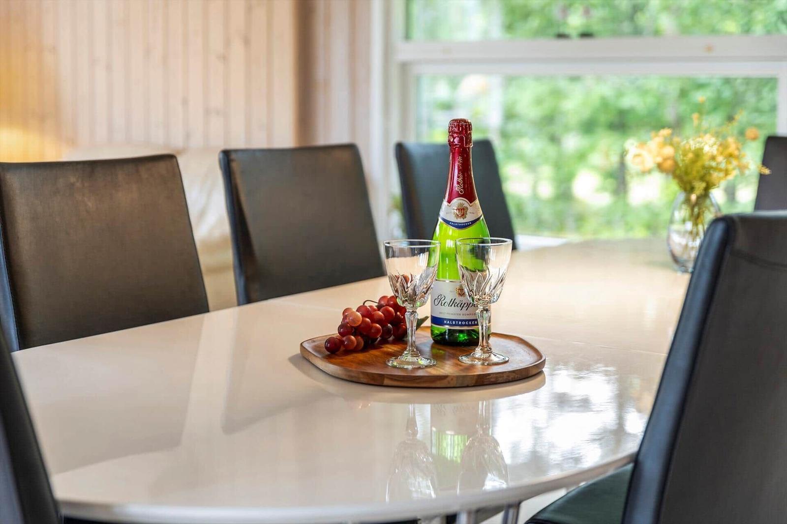 Esstisch mit Flasche, Gläsern und Trauben auf Holztablett. Fensterblick auf grüne Bäume.