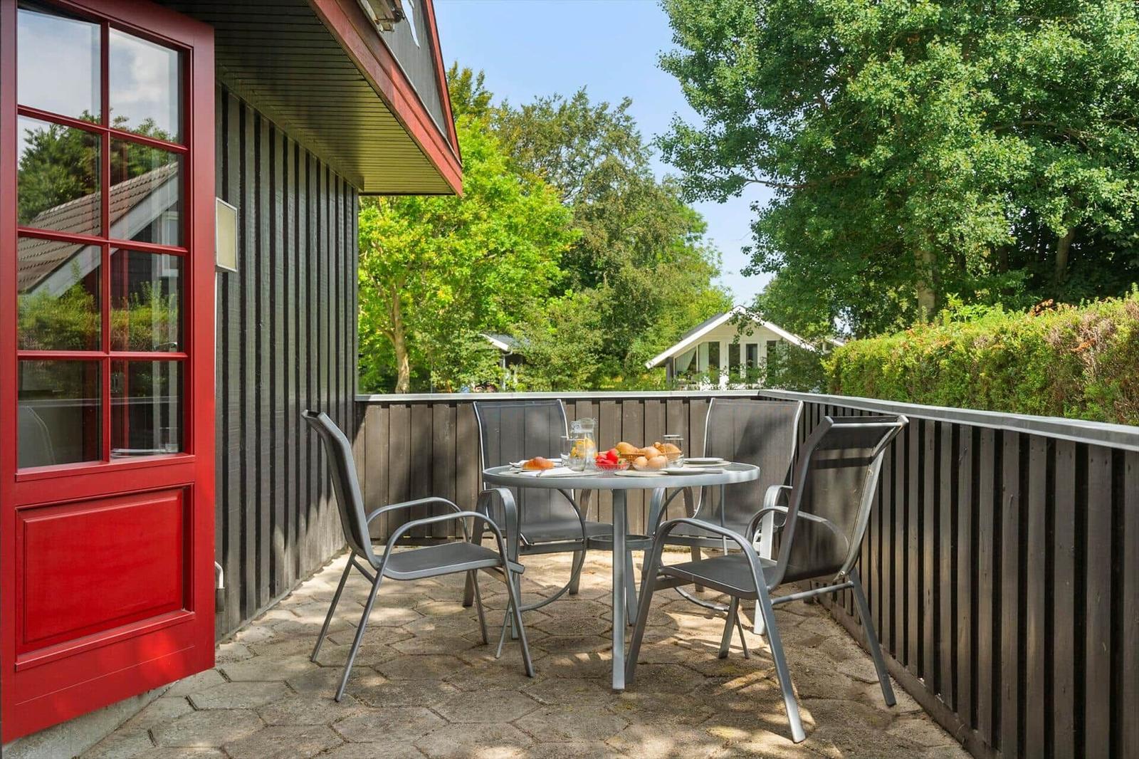 Terrace with table and chairs, red door, wooden railing, green trees in the background.