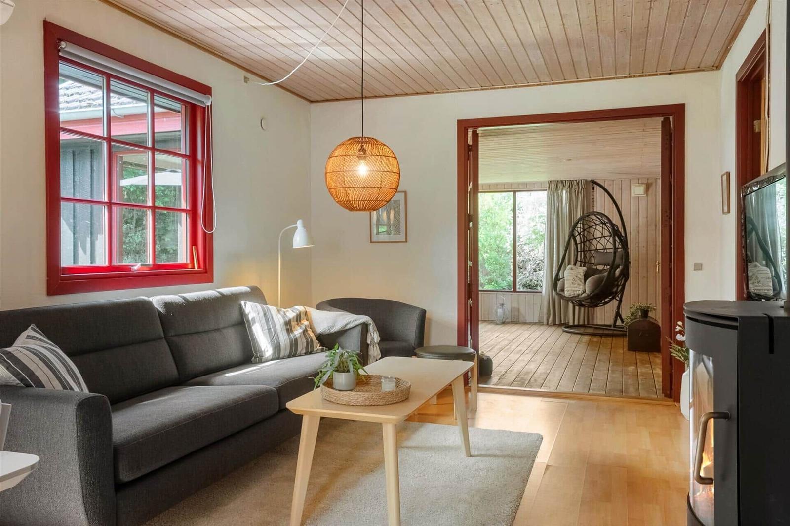 Living room with gray sofa, red window, and wooden ceiling. Entrance to terrace with hanging chair.